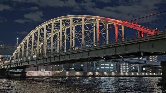 The vibrant nightlife of Tokyo featuring a commuter train crossing an illuminated arched bridge over the Sumida River, with colorful light reflections on the water.