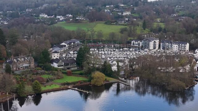An elliptical drone orbit over a large pentagon-shaped marina at Bowness. The drone turns inward as the camera tilts down, ending focused on boats moored on central pontoons with cloud reflections.