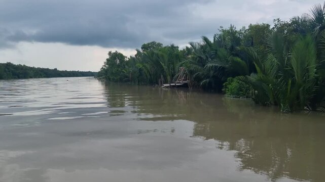 Passenger perspective from a boat slowly moving upstream the Sundarbans river in Bangladesh, with dark cloudy skies, lush riverbank vegetation, and high tide reflecting the dense mangrove surroundings
