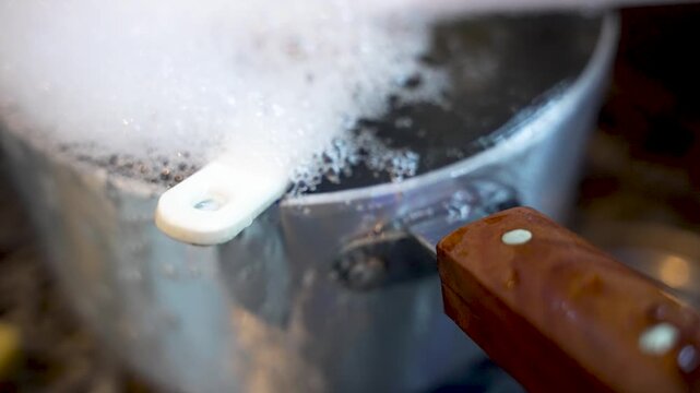 A silver pot sits in a kitchen sink overflowing with soapy water and bubbles with water pouring down the side.