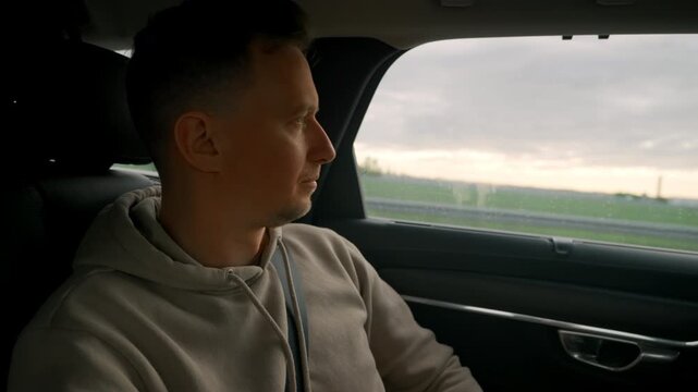 Male Passenger Gazing Out Window At Passing Fields And Sunset, Calm Expression, Seatbelt On And Hoodie