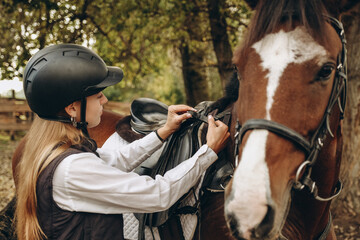 A young woman jockey adjusts the harness on her horse before training.