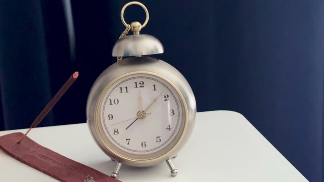 A timelapse video of a classic silver retro alarm clock, capturing the clock hands spinning rapidly. It sits on a white table against a dramatic blue background with smoke swirling in the air, creatin