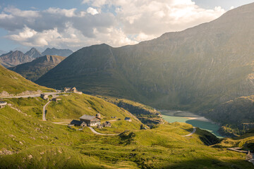 Scenic alpine mountain valley road leading toward the Pasterze Glacier with winding curves, remote mountain huts, and a turquoise glacial lake surrounded by green slopes and dramatic high Alpine peaks