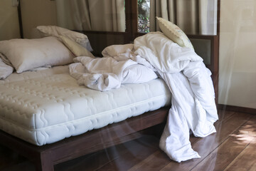 White messy bed with unmade duvet and white pillow on wooden bed frame in bedroom showing lazy or relaxed morning atmosphere with sunlight shining on floor