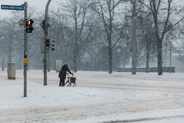 Senior woman crosses snowy city intersection with walker during winter snowfall and icy road conditions. Traffic lights and low visibility emphasize vulnerability, limited mobility and need for care