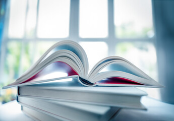 Shallow focus on pile of books and wide open book in a home pleasant atmosphere with a light widow at a unfocused background
