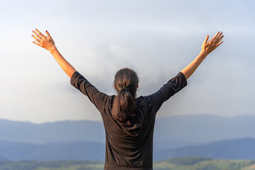 A woman stands with her arms raised toward the sky while looking out over a vast mountain range at sunset.