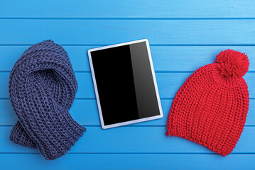 A top-down view of a digital tablet with a black screen placed between a navy blue knitted scarf and a red pom-pom beanie on a blue wooden surface.