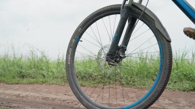 front wheel turning on dirt path, blue mountain bike with suspension fork, spinning spokes and disc brake, lowangle shot, sandals on pedals, grassy roadside, cloudy sky, rough gravel texture, active