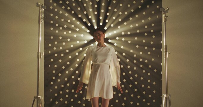 A serious woman poses against the background of a black perforated cyclorama through which light is shining