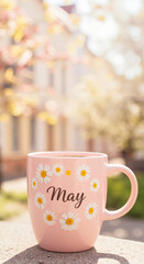Pink mug with daisies and 'May' inscription outdoors in spring  