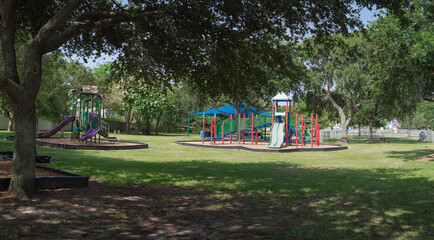 Panorama view slides and green beams contrast red-green climbers with light green slide under blue canopy. Wood chip flooring and grassy borders frame this tree-lined park in Galveston, Texas