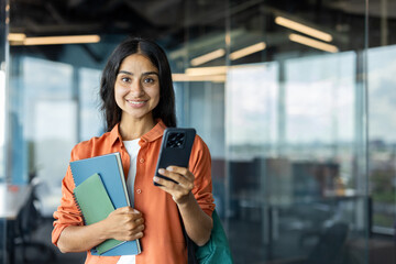 Portrait of a young Indian woman standing indoors, holding books and a mobile phone, looking at the...