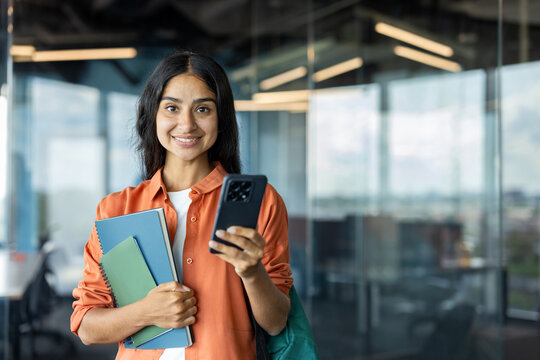 Portrait of a young Indian woman standing indoors, holding books and a mobile phone, looking at the camera