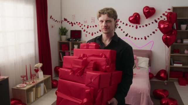 Zoom in portrait of Caucasian confident young man in black shirt posing for camera with stack of gift boxes wrapped in red paper in festive bedroom