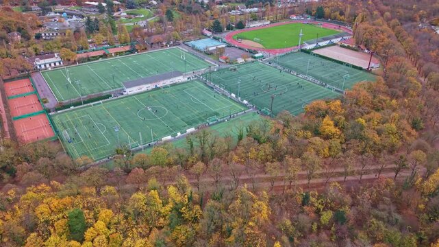 Aerial drone view of a sports complex with football, rugby, and tennis fields. A forest with vibrant autumn colors borders the facility. Ideal for sports, recreation, and seasonal concepts