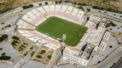 Aerial view of a large football stadium on a sunny morning. The pitch features a green pitch surrounded by white and red seating and grandstand. It is the soccer stadium of Messina, in Sicily, Italy.
