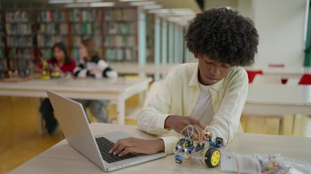 A smart African boy with curly hair building a robotic car while using a laptop to set up or write control code for operating the technology in an after-school library