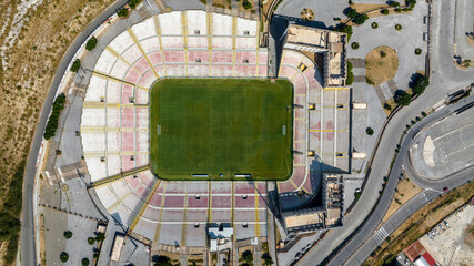 Aerial perpendicular view of a large football stadium on a sunny morning. The pitch features a green pitch surrounded by white and red seating. It is the soccer stadium of Messina, in Sicily, Italy.
