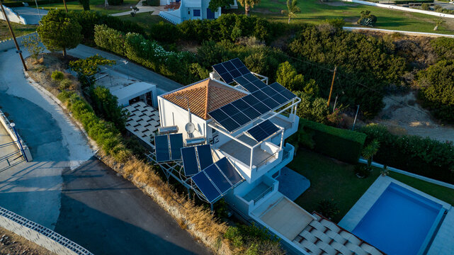 Aerial view of sun-drenched solar panels adorn the terracotta roof of a pristine white villa, casting shadows on the turquoise pool, Rhodes, Greece.