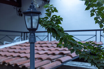 Classic black street lamp post standing next to a traditional terracotta tiled roof. Lush green foliage of a climbing plant in a Mediterranean courtyard. September, Turkey.