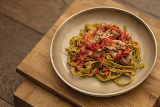 Exquisite fine dining tagliatelle pasta tossed in vibrant pesto, topped with tender grilled chicken and a zesty tomato concass&eacute;. Elegantly served on a rustic wooden table in a luxury bistro.