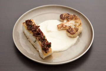A gourmet fine dining dish featuring pan-seared white fish topped with olive tapenade, served over a silky cauliflower pur&eacute;e and charred cauliflower steak. Elegant, minimalist culinary plating.