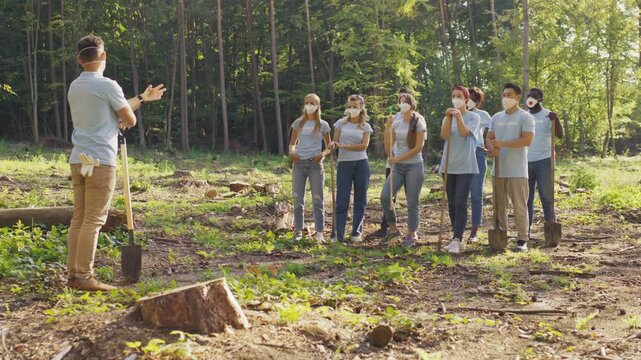 Wide forest scene showing masked volunteer group standing apart. Men with women holding shovels and tools. Caucasian male coordinator explaining safety rules. Preparing for eco work.