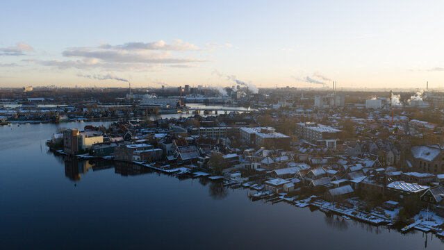 Aerial view of a town dusted with snow along the serene waterfront, with industrial structures releasing smoke into the pale sky, Zaandam, Netherlands.