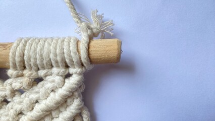 Macrame craft detail of cream colored cotton rope tied to a wooden dowel, showing the texture of handmade knots against a white background. © RiyanBudi