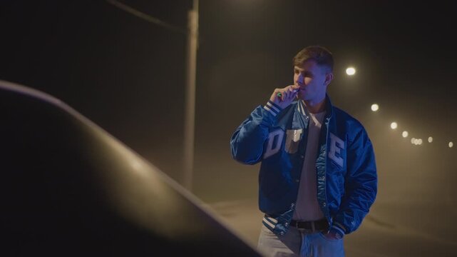 night roadside white man by car leaning on hood, blue jacket gleaming from headlight reflections, vaping and scanning dark highway, cinematic tension of waiting driver, possible courier on pause,