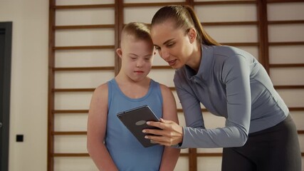 Supportive female trainer helping boy with down syndrome use digital tablet during inclusive sports class promoting learning, physical activity and confidence