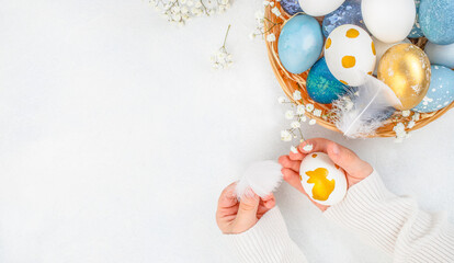 Hands holding painted Easter egg near basket with blue and gold eggs on white background with copy space