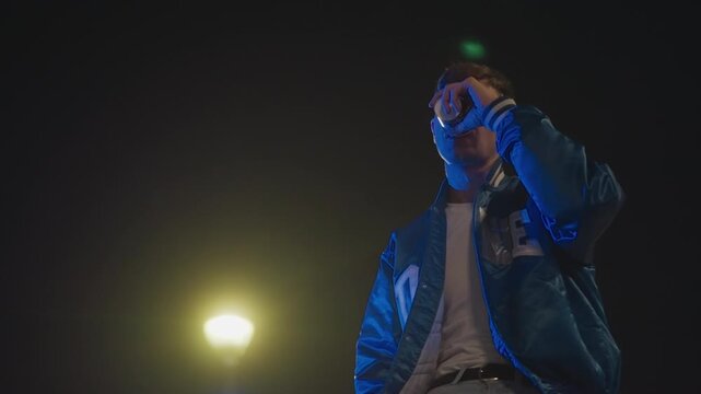young white man under streetlight, leaning by parked car in neonlit lot wearing blue varsity bomber jacket, contemplative gaze to sky, cigarette in hand, moody cinematic grain, urban solitude