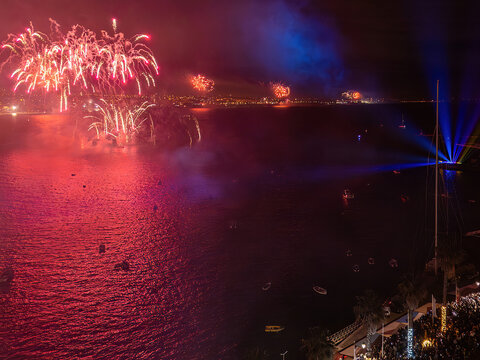 Aerial view of vibrant fireworks exploding over the dark ocean, contrasting with the illuminated shoreline and boats, Cascais, Lisbon, Portugal.