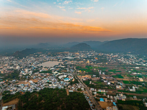 Aerial view of a sprawling town nestled amidst rolling hills, where structures meet open fields under a soft, golden sky, Pushkar Rural, Rajasthan, India.