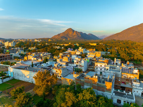 Aerial view of the sun-kissed rooftops contrasting against the rugged Aravalli hills, bathed in the warm glow of the setting sun, Pushkar Rural, Rajasthan, India.