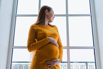 Pregnancy motherhood people expectation future. Pregnant woman with big belly standing near window at home. Girl hugging her tummy enjoying pregnancy. Maternity tenderness parenthood new life concept
