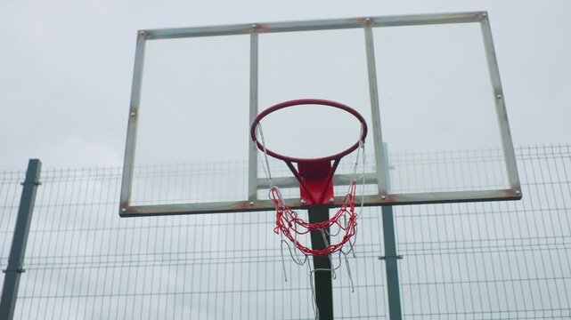 Centered backboard with red rim, empty outdoor court and chainlink fence symmetrical composition and subtle rust on frame create calm minimal mood, ideal for sports background, architectural