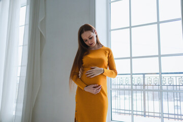 Pregnancy motherhood people expectation future. Pregnant woman with big belly standing near window at home. Girl hugging her tummy enjoying pregnancy. Maternity tenderness parenthood new life concept