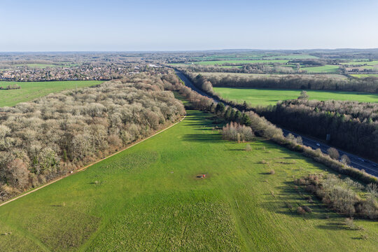 Aerial view of a vibrant green field juxtaposed against the stark contrast of a dense forest alongside a long highway, Basingstoke, England, United Kingdom.