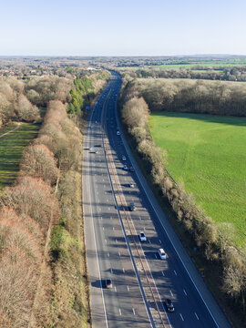 Aerial view of vehicles glide along the grey motorway, flanked by bare trees and vibrant green fields, a stark contrast of natural and man-made landscapes, Basingstoke, England, United Kingdom.