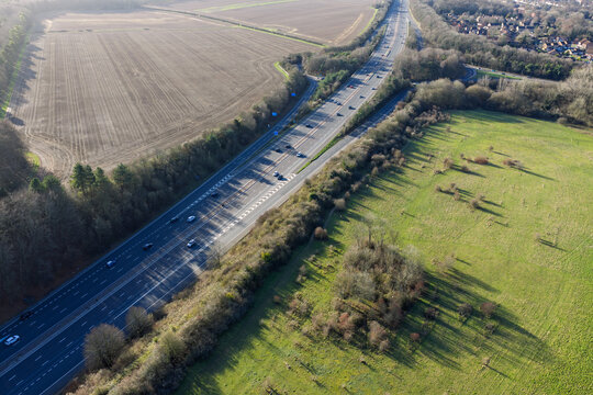 Aerial view of a long highway cutting through fields and forests, the dark asphalt contrasting with the bright green grass, Basingstoke, England, United Kingdom.