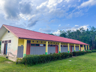 A vibrant Indonesian rural school building with a colorful exterior and red roof, set against a cloudy blue sky and surrounded by trees and grass, embodies the spirit of education.