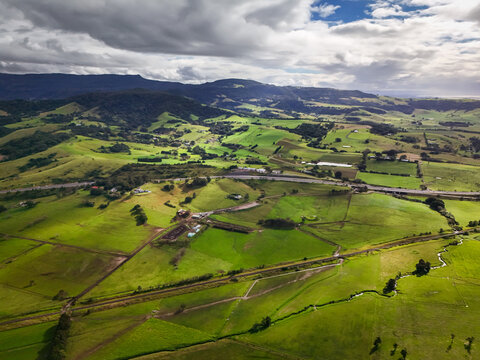 Aerial view of lush green fields meet the distant, hazy mountains under a sky streaked with clouds, Crooked River Road, Gerroa, New South Wales, Australia.