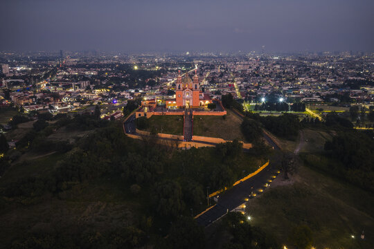 Aerial view of the illuminated Great Pyramid of Cholula crowned by the Templo de la Virgen de los Remedios against the fading twilight, Cholula, Puebla, Mexico.