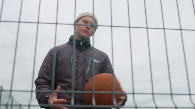 young white male with basketball behind fence, standing on outdoor court in beanie and jacket, holding ball with confident yet pensive look, overcast sky, metal grid foreground, urban streetball vibe,
