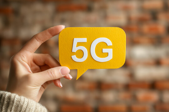Hand holding a yellow speech bubble cutout with white lettering symbolizing advanced fifth generation wireless network technology on a blurred wall background - Powered by Adobe