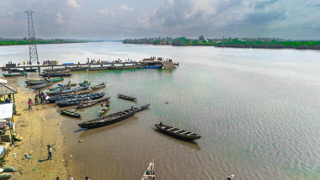 Aerial view of boats clustered along the sandy shore and a jetty extending into the calm river under a cloudy sky, Khana, Bori, Nigeria.